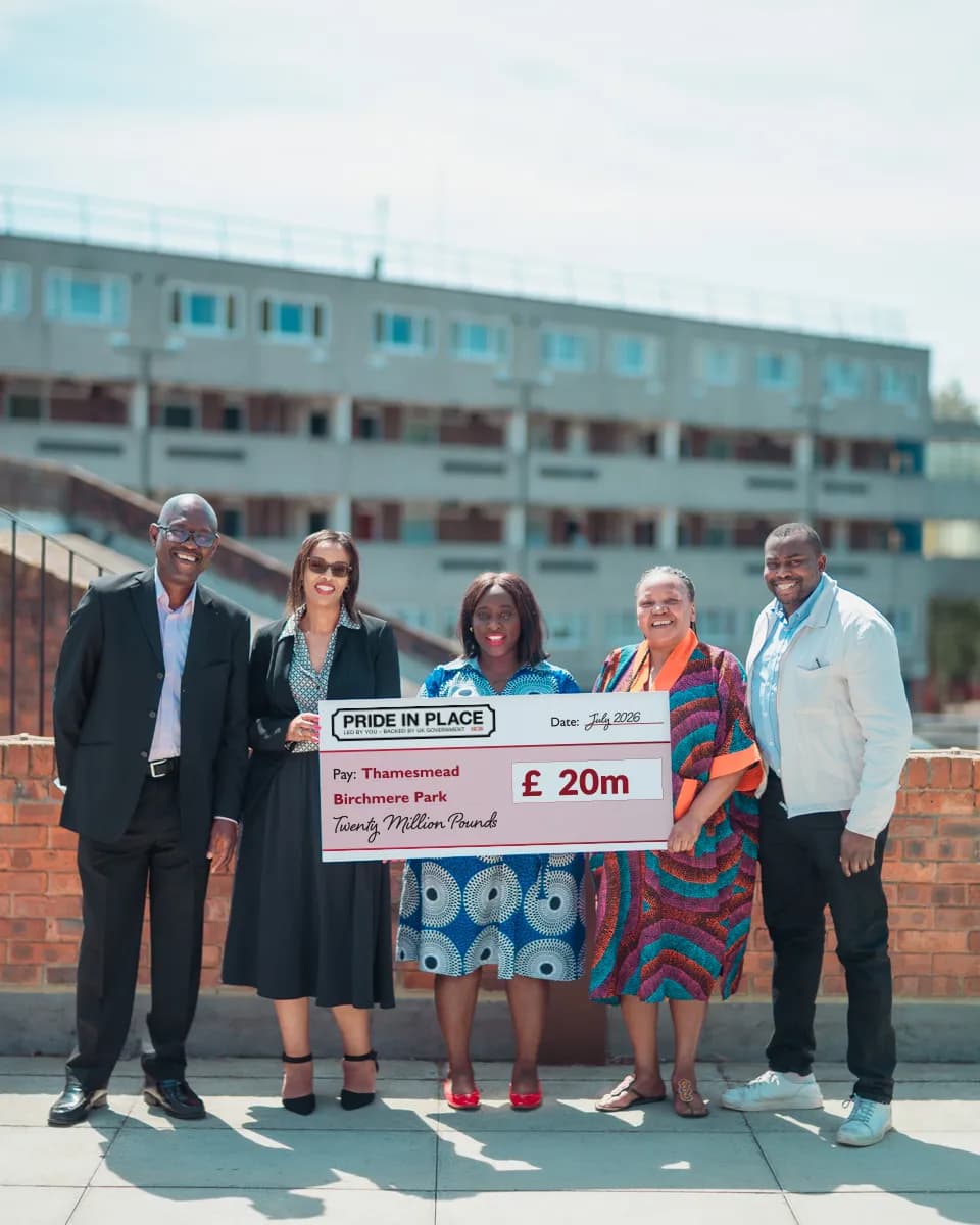Five adults, three women and two men, stand smiling on a rooftop with a large novelty check for Β£20 million, with a residential building in the background.